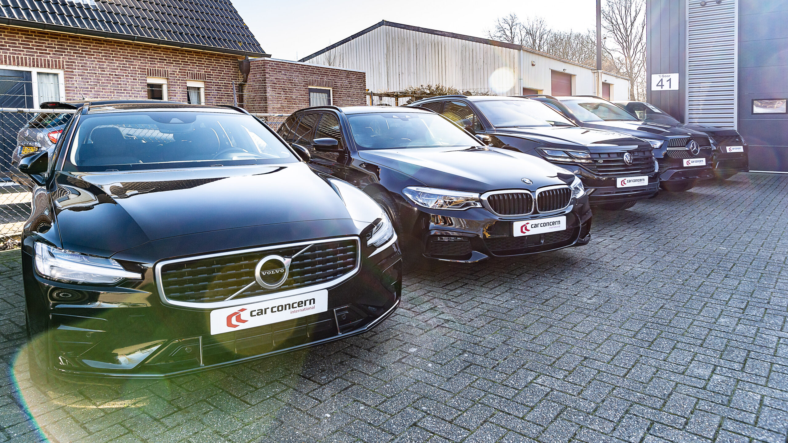 Zwarte Volvo V60, BMW 5 Serie Touring, Volkswagen Touareg en Mercedes-Benz GLE in een rij voor het pand